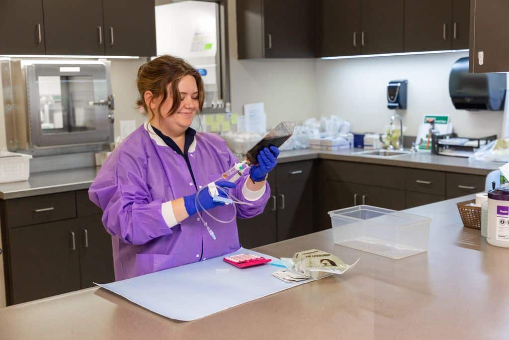 Lab technician in a purple protective gown and gloves preparing an IV medication bag at the prep counter at Gadsden Cancer Clinic