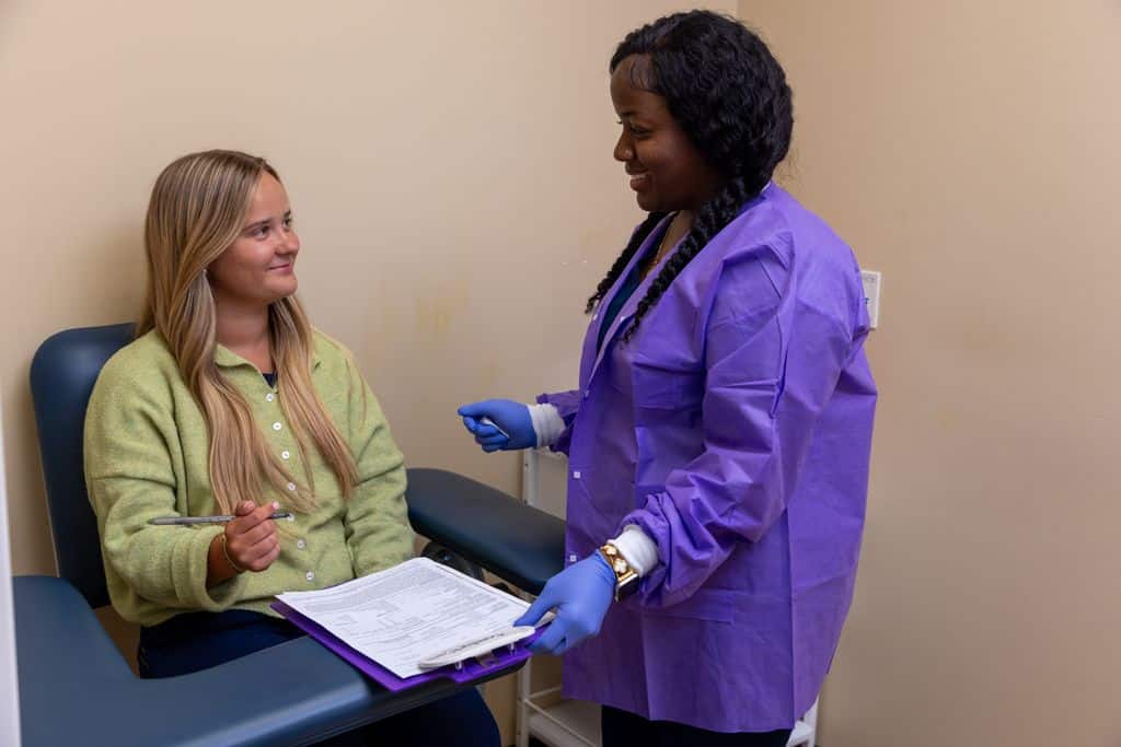 Staff member in a purple protective gown assisting a seated patient with paperwork in an exam room at Gadsden Cancer Clinic