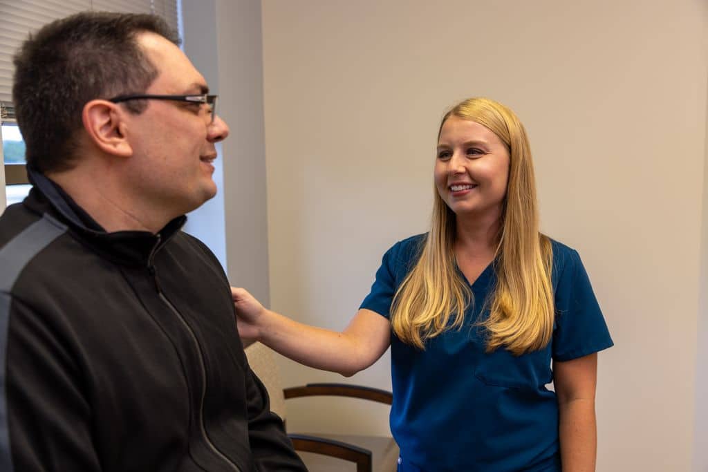 Nurse in blue scrubs resting a reassuring hand on a standing patient's shoulder in an exam room at Gadsden Cancer Clinic