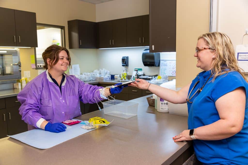 Clinician in a purple gown and blue gloves handing treatment supplies to a colleague in scrubs at a prep counter at Gadsden Cancer Clinic